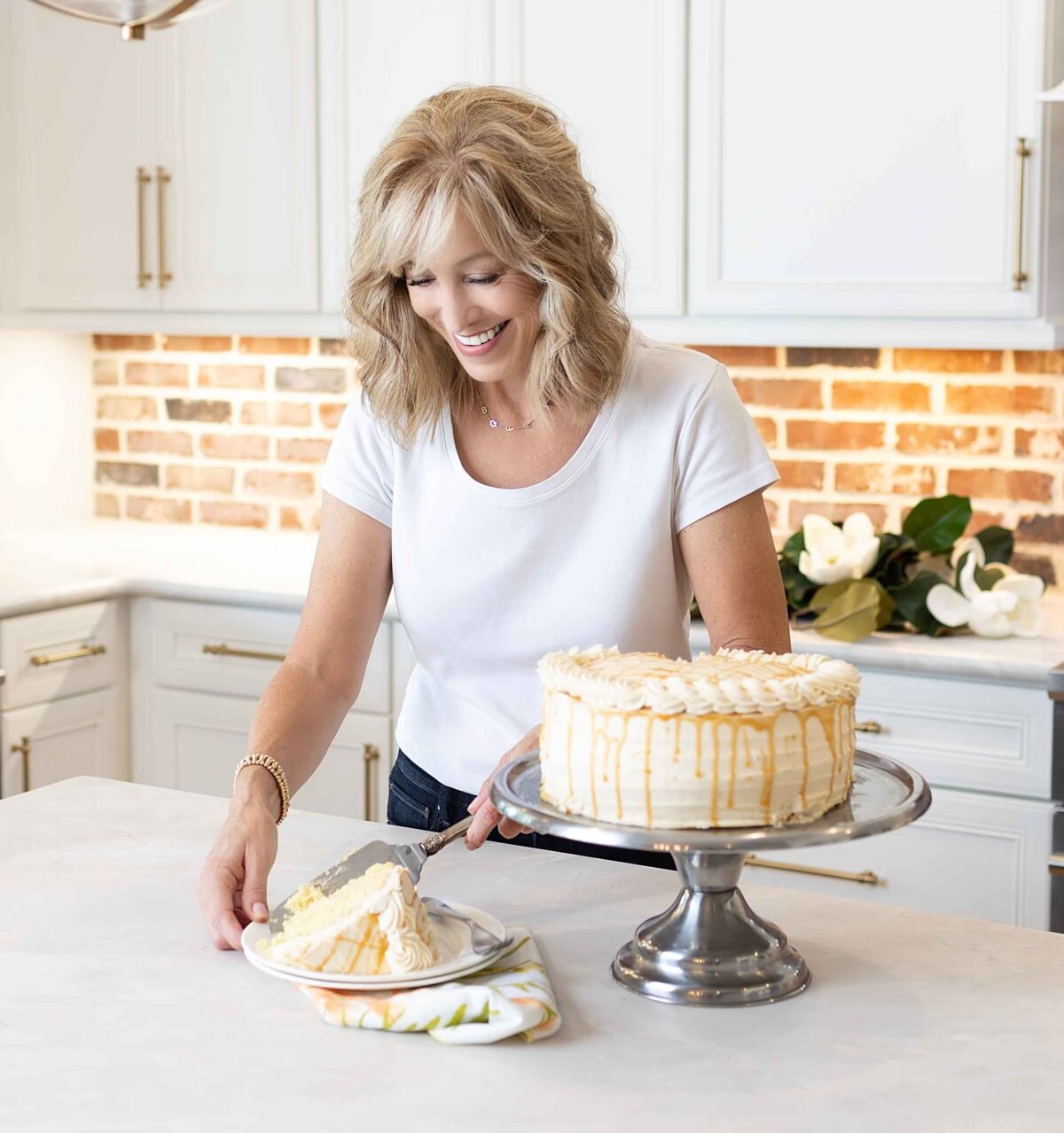 A woman in a white shirt is cutting a slice of a cream-colored cake with drizzled topping in a modern kitchen.