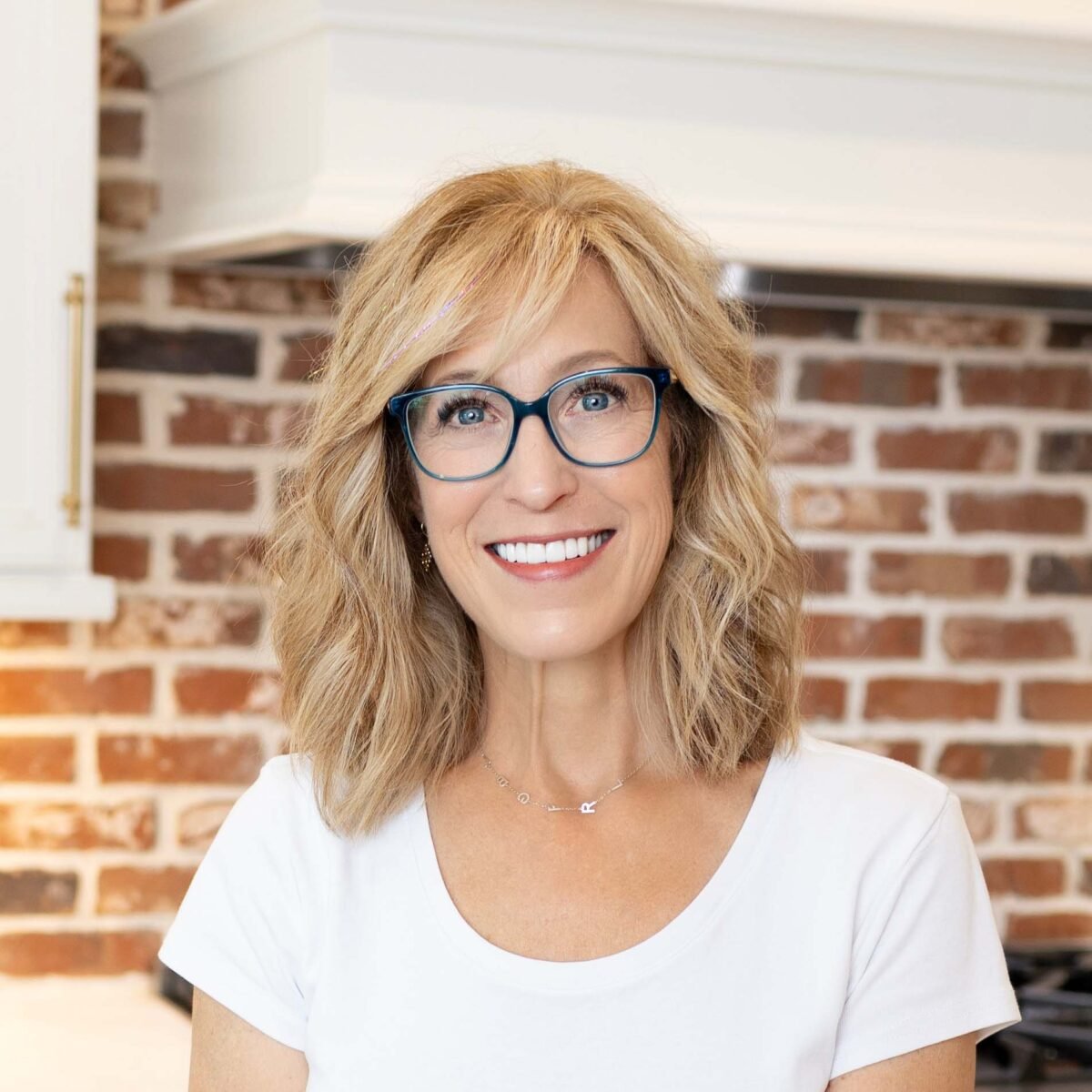 Person with shoulder-length blond hair and blue glasses smiling indoors in front of a red brick wall.