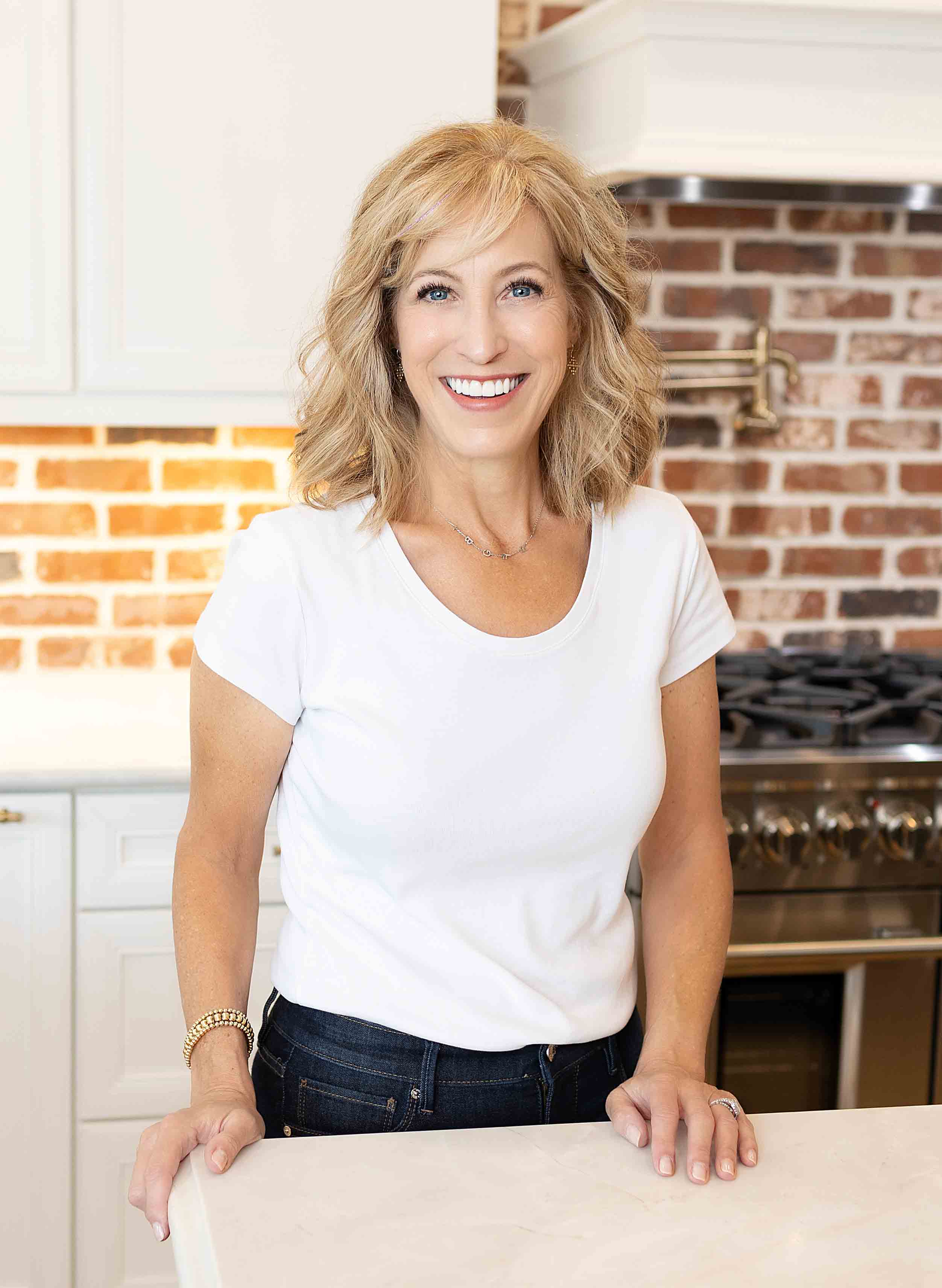 A woman with shoulder-length blonde hair, wearing a white T-shirt and dark jeans, stands in a kitchen with a brick backsplash and stainless steel appliances. She is smiling and resting her hands on the counter.