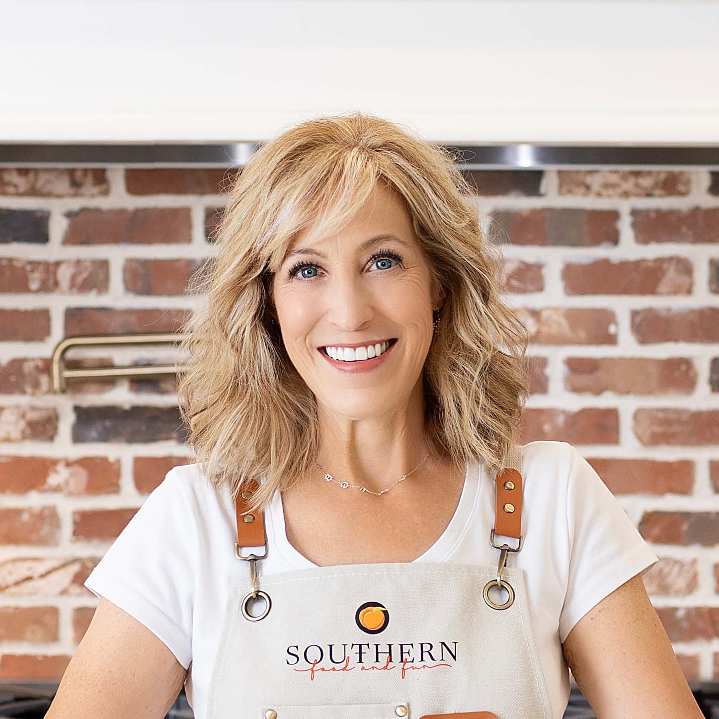 Woman with blonde hair wearing a white shirt and an apron with a logo that reads "Southern" stands in a kitchen with a brick backsplash. She is smiling and looking at the camera.