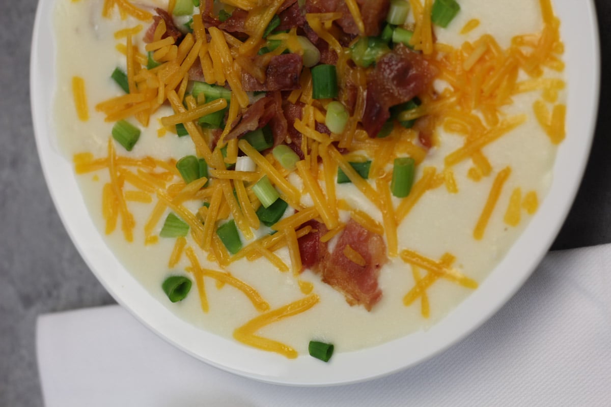 A bowl of creamy potato soup topped with shredded cheddar cheese, chopped green onions, and crumbled bacon, placed on a white cloth napkin.