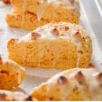 Close-up of triangular scones with a golden-brown crust and topped with a white glaze, arranged on a white surface.