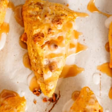 Close-up of freshly baked, triangular pumpkin scones with white glaze and caramel drizzle on a light-colored surface.