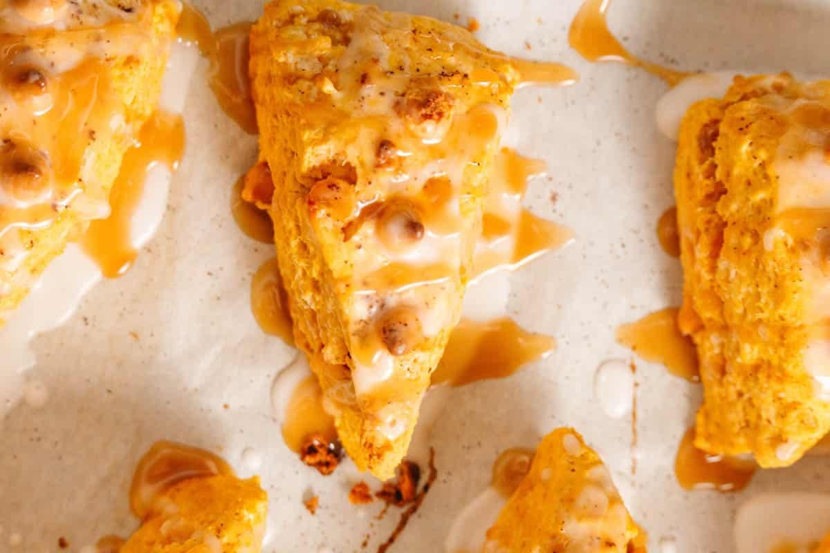 Close-up of freshly baked, triangular pumpkin scones with white glaze and caramel drizzle on a light-colored surface.