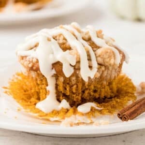 A partially unwrapped muffin with crumb topping and white icing drizzled on top sits on a plate next to a small cinnamon stick.