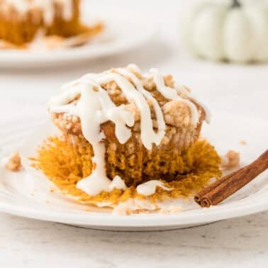 A pumpkin streusel muffin with white icing on a plate, partially unwrapped with a cinnamon stick beside it. Another dessert and decorative pumpkins are blurred in the background.