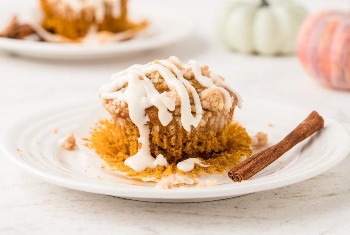 A pumpkin streusel muffin with white icing on a plate, partially unwrapped with a cinnamon stick beside it. Another dessert and decorative pumpkins are blurred in the background.