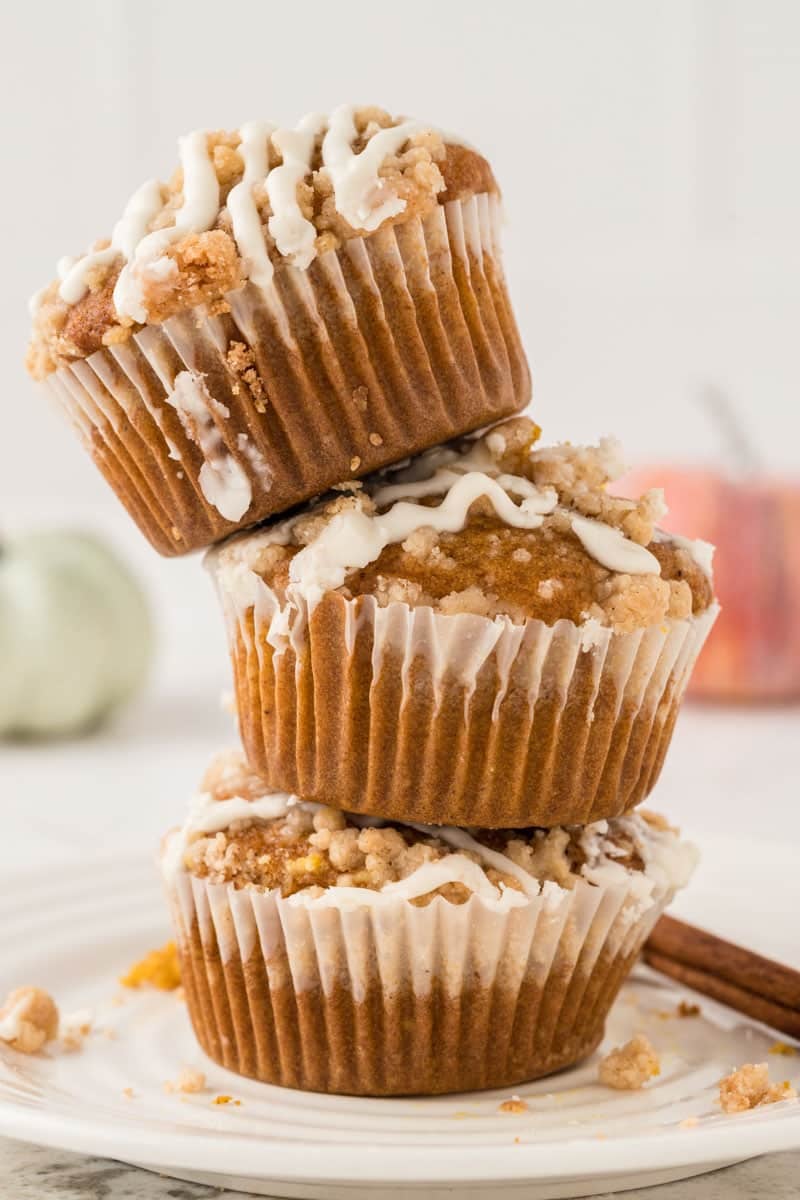 A stack of three pumpkin muffins topped with white icing, placed on a white plate. A cinnamon stick lies beside the muffins. The background is softly blurred with a hint of a pumpkin.