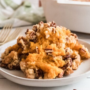 A serving of sweet potato casserole topped with a pecan and crumb mixture sits on a white plate, with a fork on the table beside it.