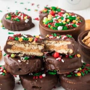 Chocolate-covered Ritz crackers with festive sprinkles and a peanut butter filling, stacked with some cut in half to show the inside. Bowls of colorful sprinkles are in the background.