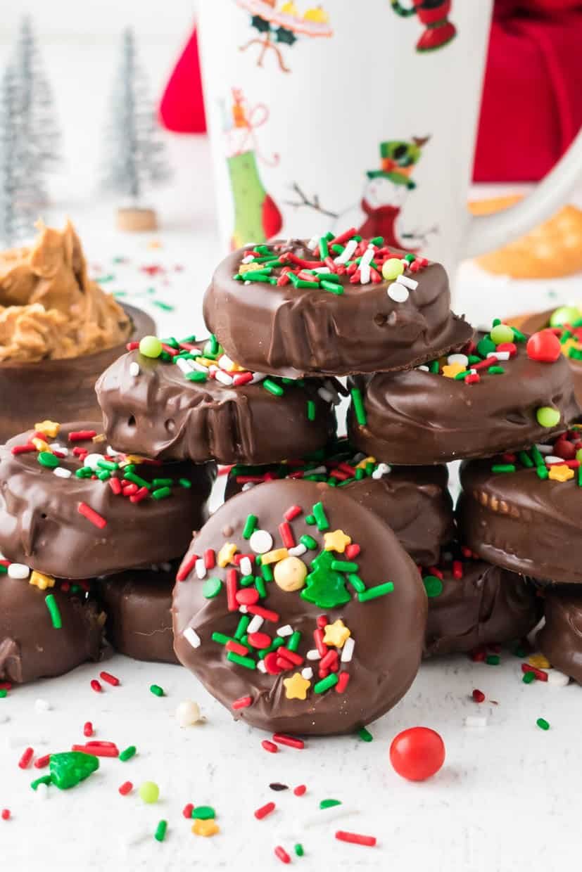 A stack of Chocolate-covered Ritz crackers decorated with colorful sprinkles and a bowl of peanut butter in the background. A festive mug and small decorative trees are also visible.