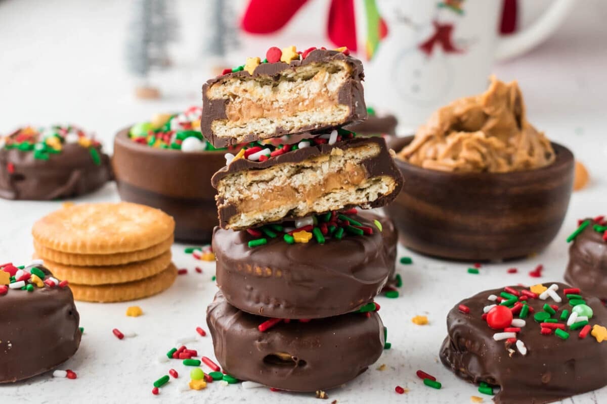 Stack of chocolate-covered peanut butter crackers topped with holiday sprinkles, with a mug, peanut butter, and plain crackers in the background.