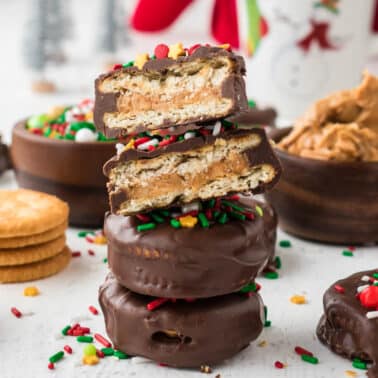Stack of chocolate-covered peanut butter crackers topped with holiday sprinkles, with a mug, peanut butter, and plain crackers in the background.