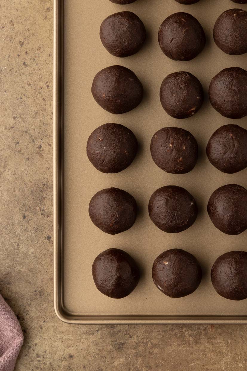 A tray of evenly spaced, round chocolate dough balls on a metal baking sheet, placed on a textured countertop.