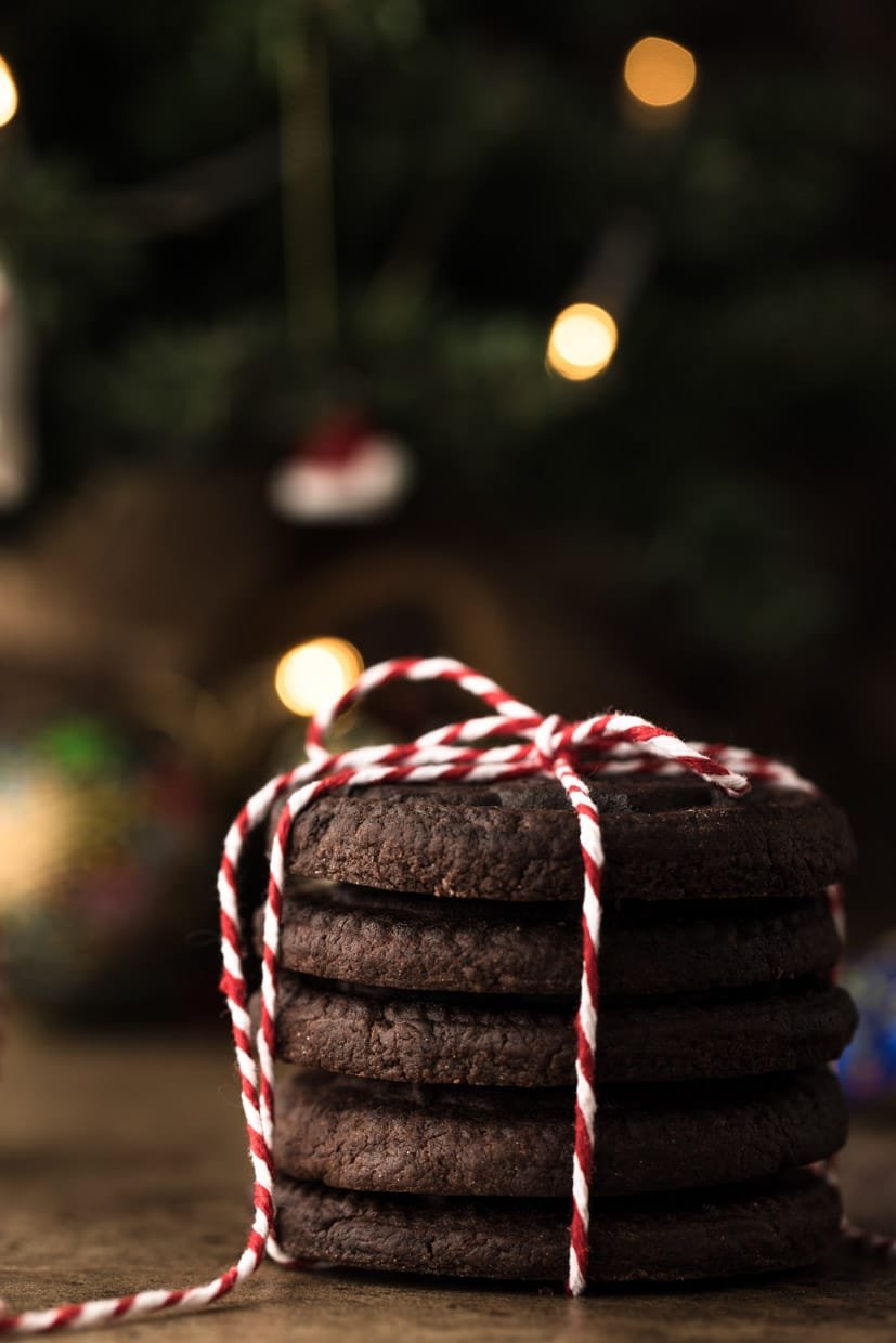 Stack of five chocolate cookies tied with red and white string, in front of blurred Christmas tree lights.