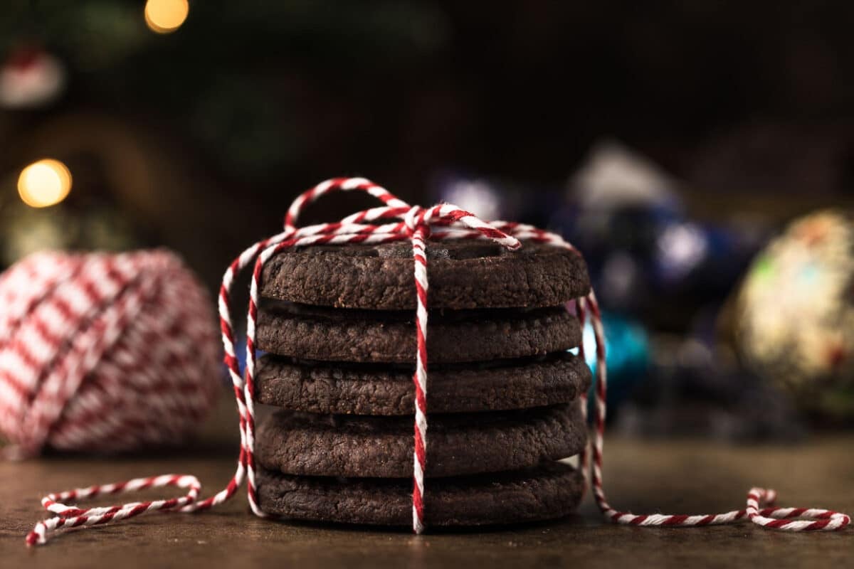 A stack of chocolate cookies tied with red and white string is placed on a table. In the background, there are blurred festive decorations.