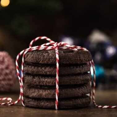 A stack of chocolate cookies tied with red and white string is placed on a table. In the background, there are blurred festive decorations.