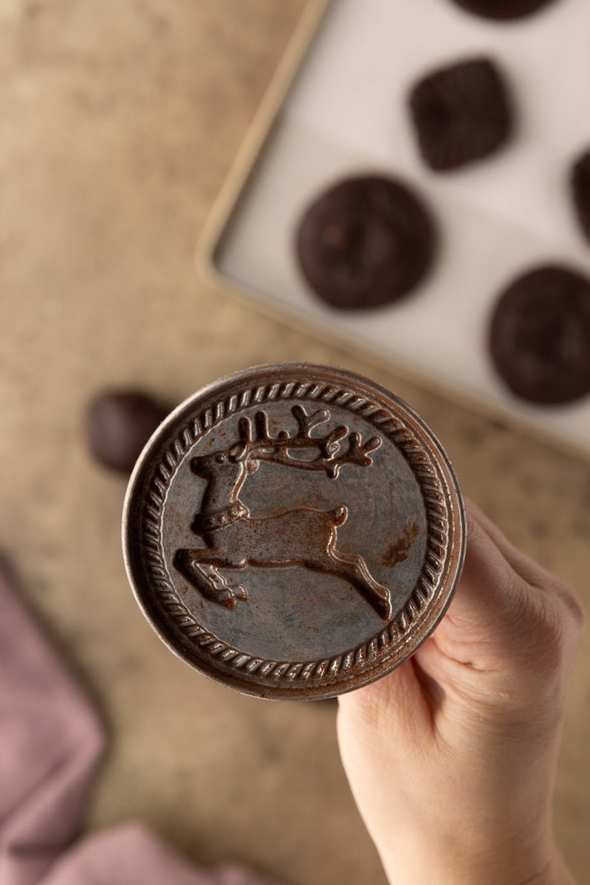 Hand holding a cookie stamp with a reindeer design; cookies are on a tray in the background.