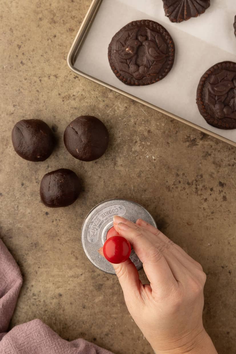 A hand presses a cookie stamp into dark dough balls on a textured surface beside a tray with stamped cookies.