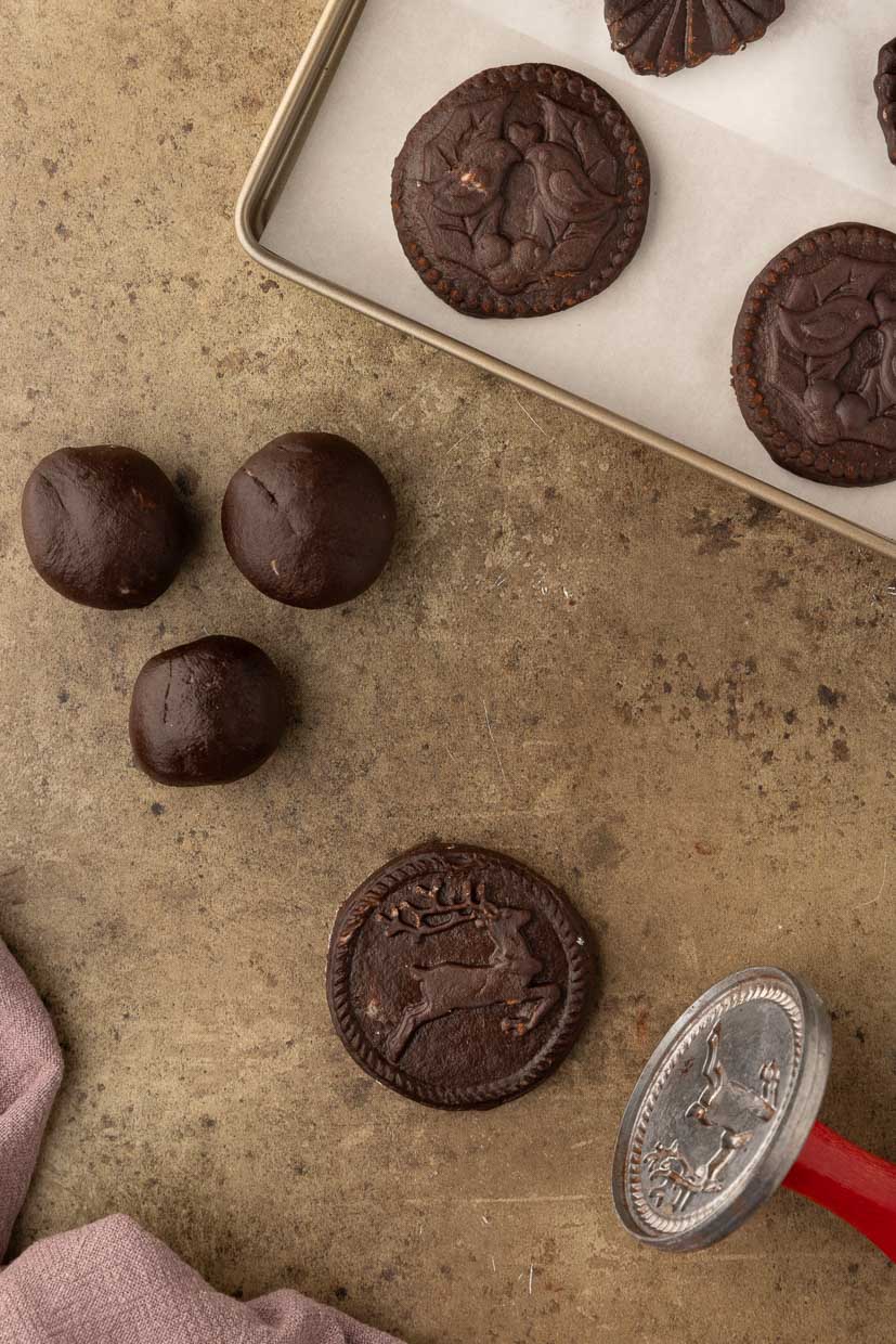 Chocolate cookies on a baking tray next to unbaked cookie dough balls and a cookie stamp with a reindeer design on a textured surface.