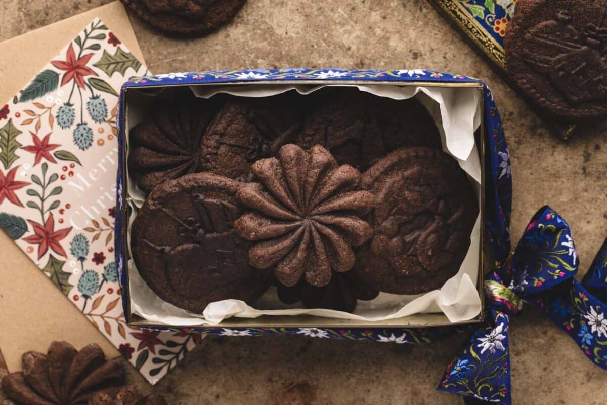 A decorative box filled with various chocolate cookies is displayed on a surface, alongside a floral card and a blue ribbon.