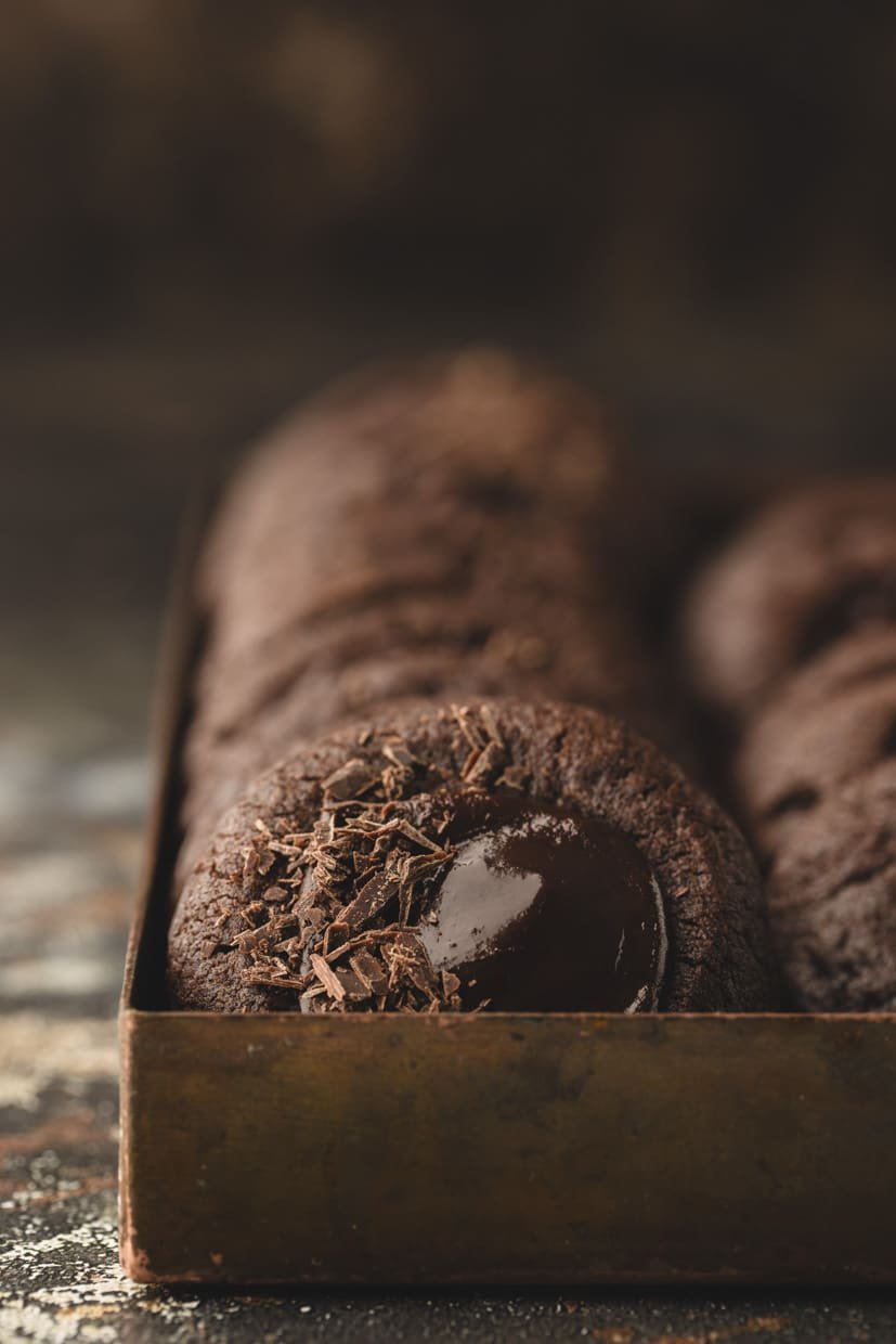 A close-up of thumbprint cookies with chocolate in a metal tray, focusing on one cookie topped with chocolate shavings and a glossy chocolate center.