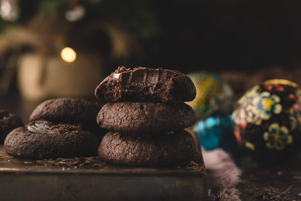 A stack of thumbprint cookies with chocolate with one on top partially eaten, set on a wooden surface. Blurry decorative ornaments are visible in the background.