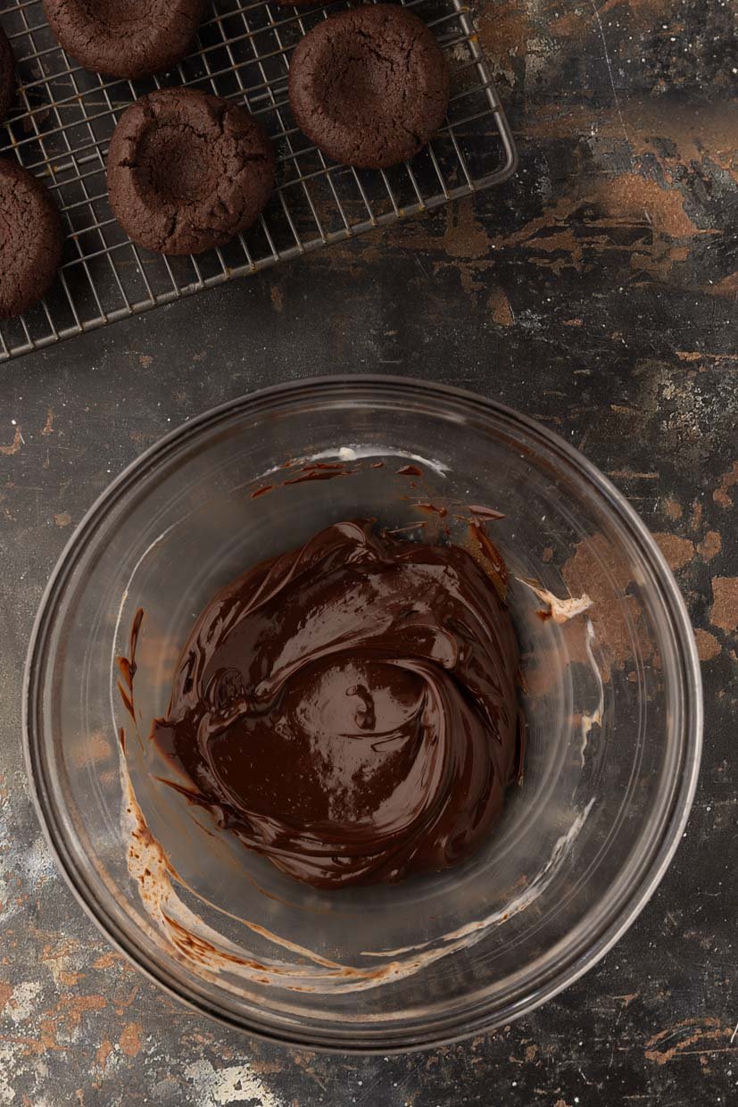 A glass bowl with melted chocolate next to a cooling rack holding several baked chocolate cookies on a textured surface.