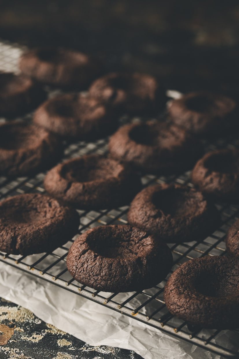 A cooling rack with multiple dark chocolate cookies, each with an indentation in the center, placed on parchment paper.