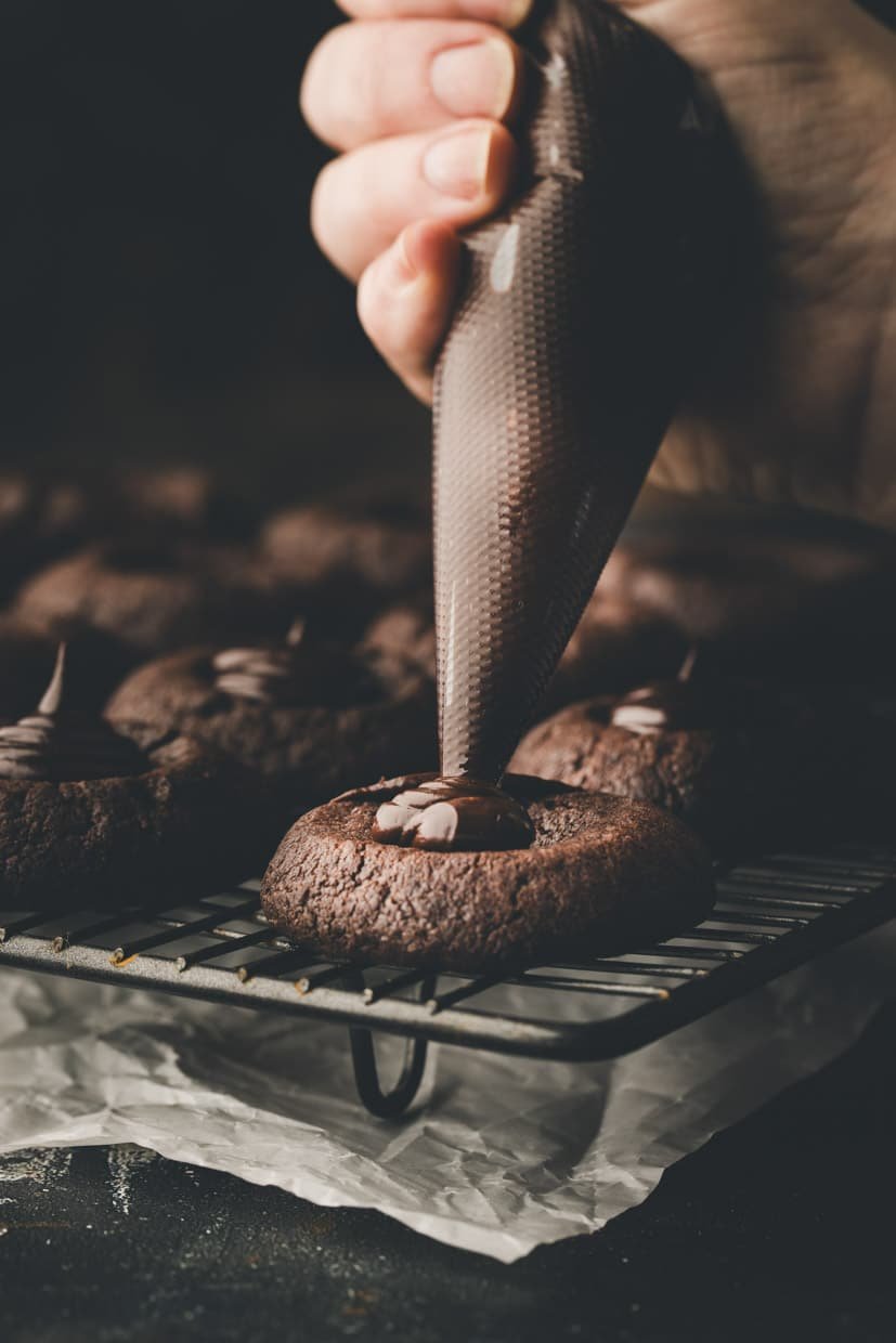 A hand piping chocolate into the center of a chocolate cookie on a cooling rack.