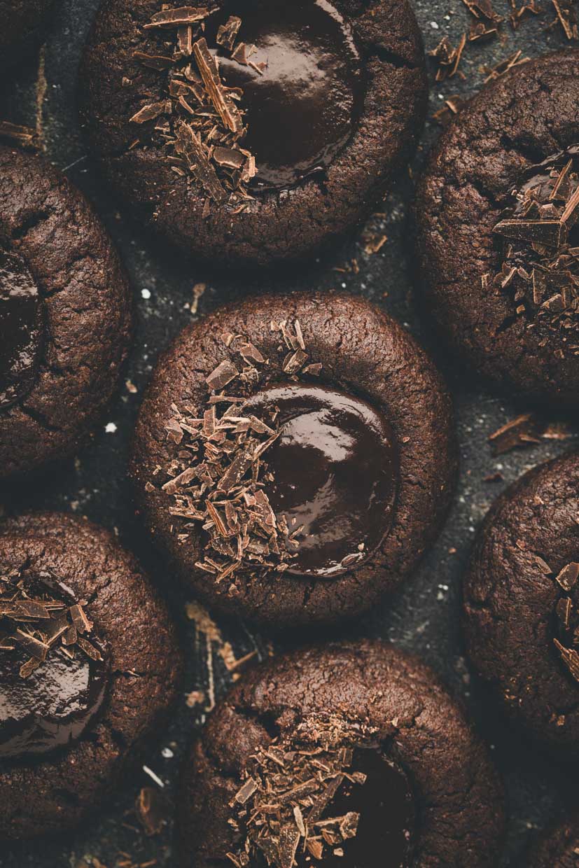 Close-up of chocolate thumbprint cookies topped with glossy chocolate and chocolate shavings, arranged on a dark surface.