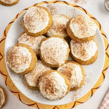 A plate of frosted cookies sprinkled with cinnamon is displayed on a table, surrounded by a few additional cookies and a metal sifter on the side.