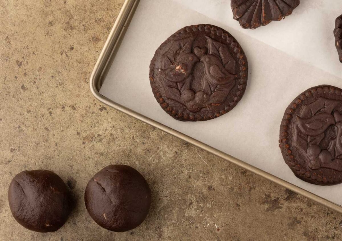 Chocolate molded cookies rest on a baking tray lined with parchment paper, sitting alongside two balls of the best cookie dough on a brown surface, ready to make cookies that delight.