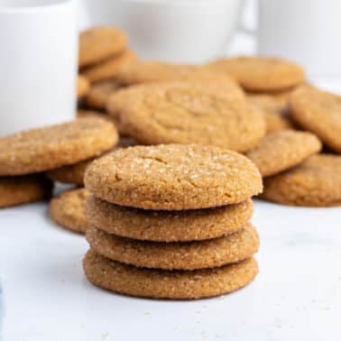 A stack of sugar-coated ginger cookies with more cookies and mugs in the background on a white surface.