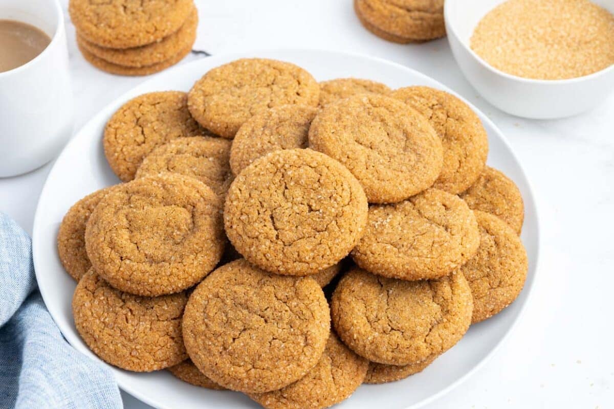 A plate of freshly baked ginger cookies is displayed on a white surface, with a cup of coffee and a bowl of sugar in the background.