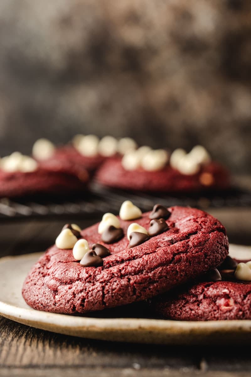 Close-up of red velvet cookies topped with white and dark chocolate chips on a plate, with more cookies in the blurred background.