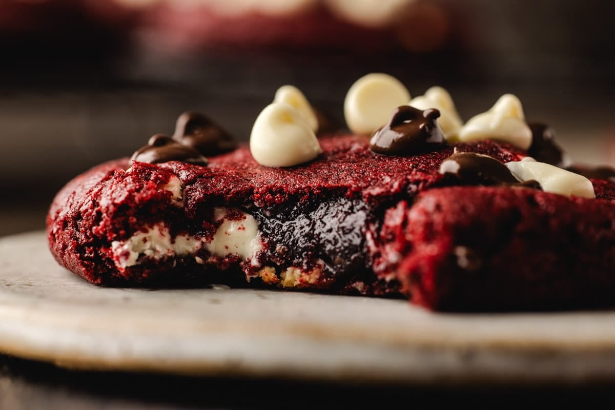 Close-up of a red velvet cookie with a bite taken, topped with white and dark chocolate chips, on a round plate.