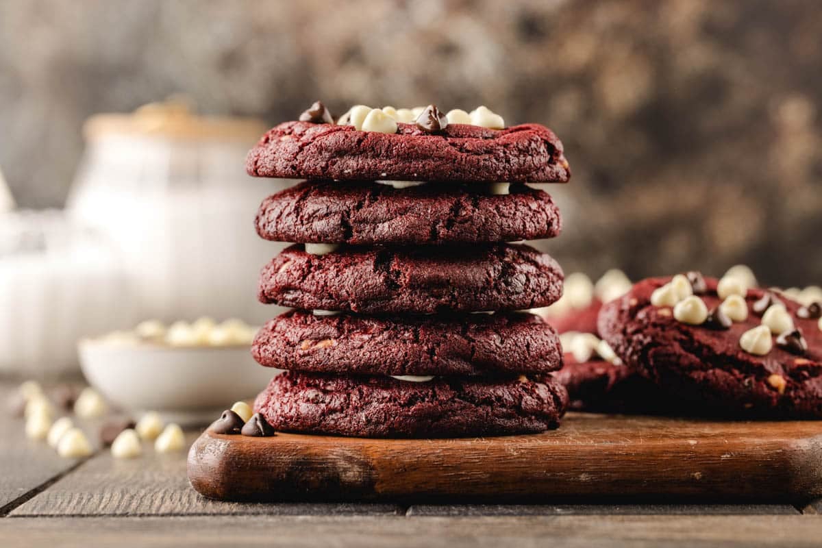 A stack of red velvet cookies with white and dark chocolate chips on a wooden board, with a jar and bowl in the blurred background.