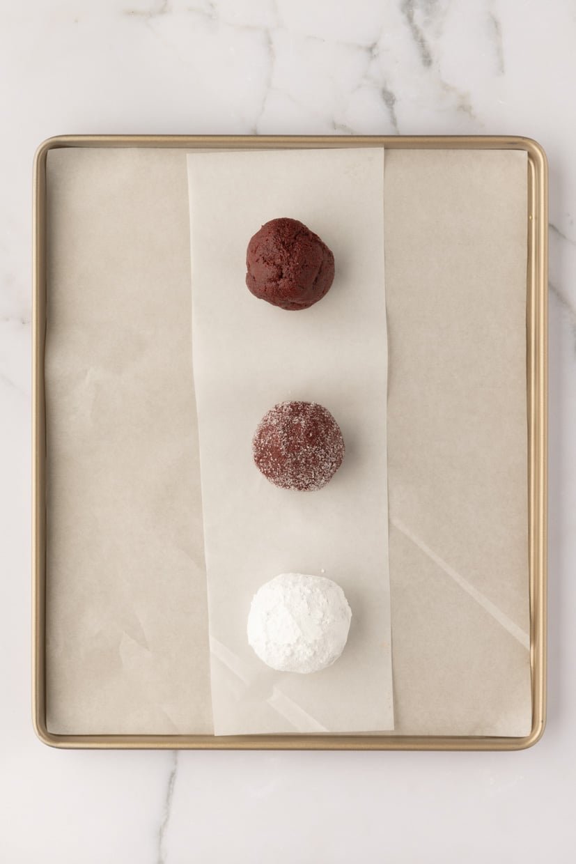 Three chocolate dough balls on a parchment-lined baking tray, with the top one plain, the middle one partially coated in sugar, and the bottom one fully coated in powdered sugar.