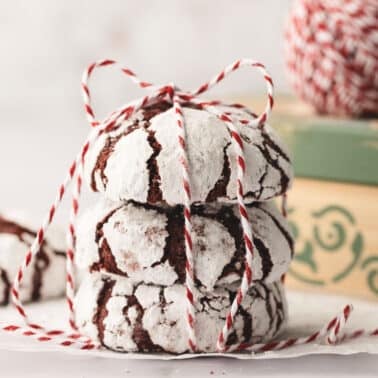 Three red velvet crinkle cookies tied with red and white string, placed on parchment paper. A ball of string and a green box are in the background.