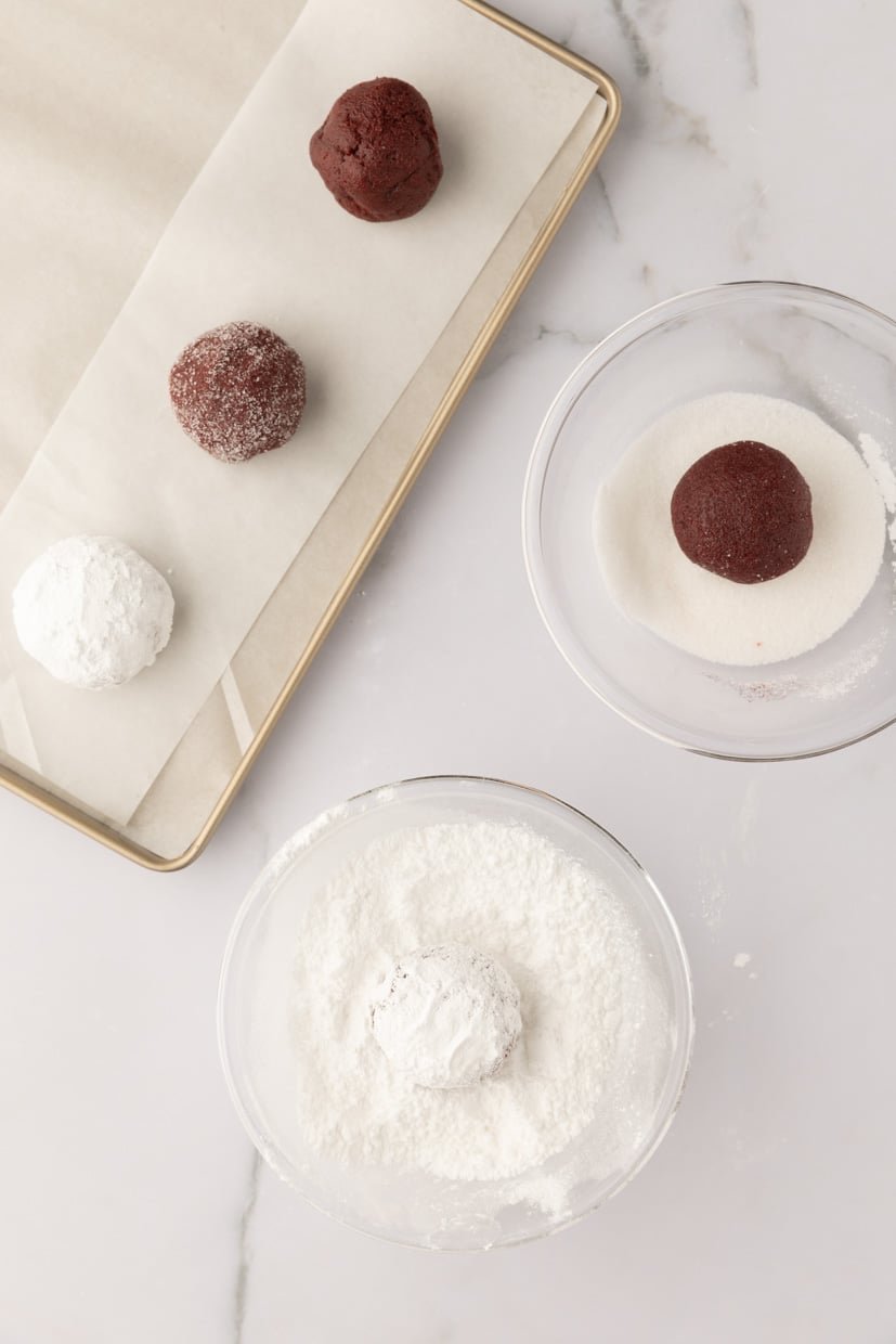 Chocolate cookie dough balls being coated in powdered sugar on a marble surface.