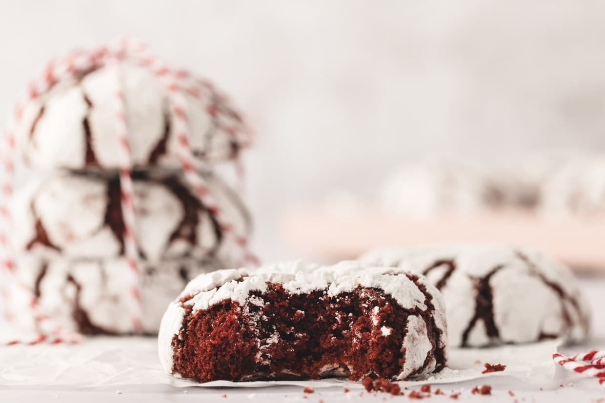 Close-up of red velvet crinkle cookies, one with a bite taken out, showing soft red interior and white powdered surface. Three whole cookies stacked in the background.