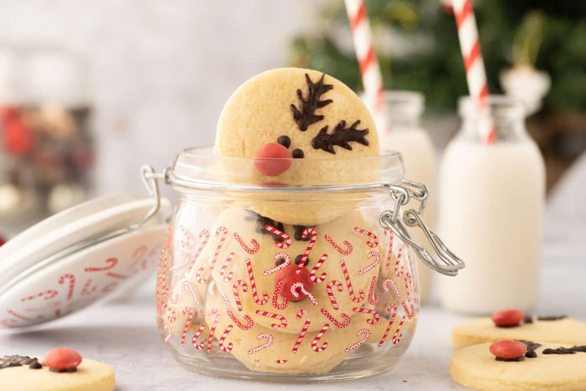 A clear jar decorated with candy cane patterns contains reindeer sugar cookies. Two bottles of milk with striped straws are visible in the background.