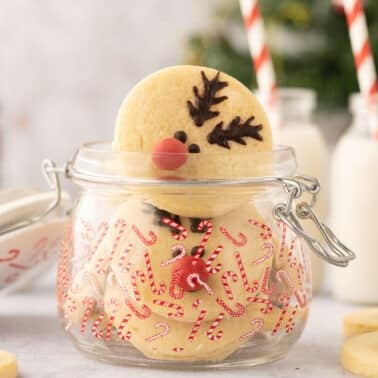 A clear jar decorated with candy cane patterns contains reindeer sugar cookies. Two bottles of milk with striped straws are visible in the background.