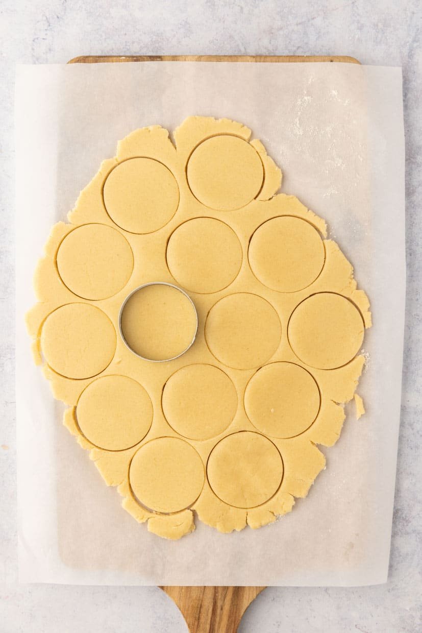 Rolled cookie dough on parchment paper with several round shapes cut out and one cookie cutter visible on a wooden board.