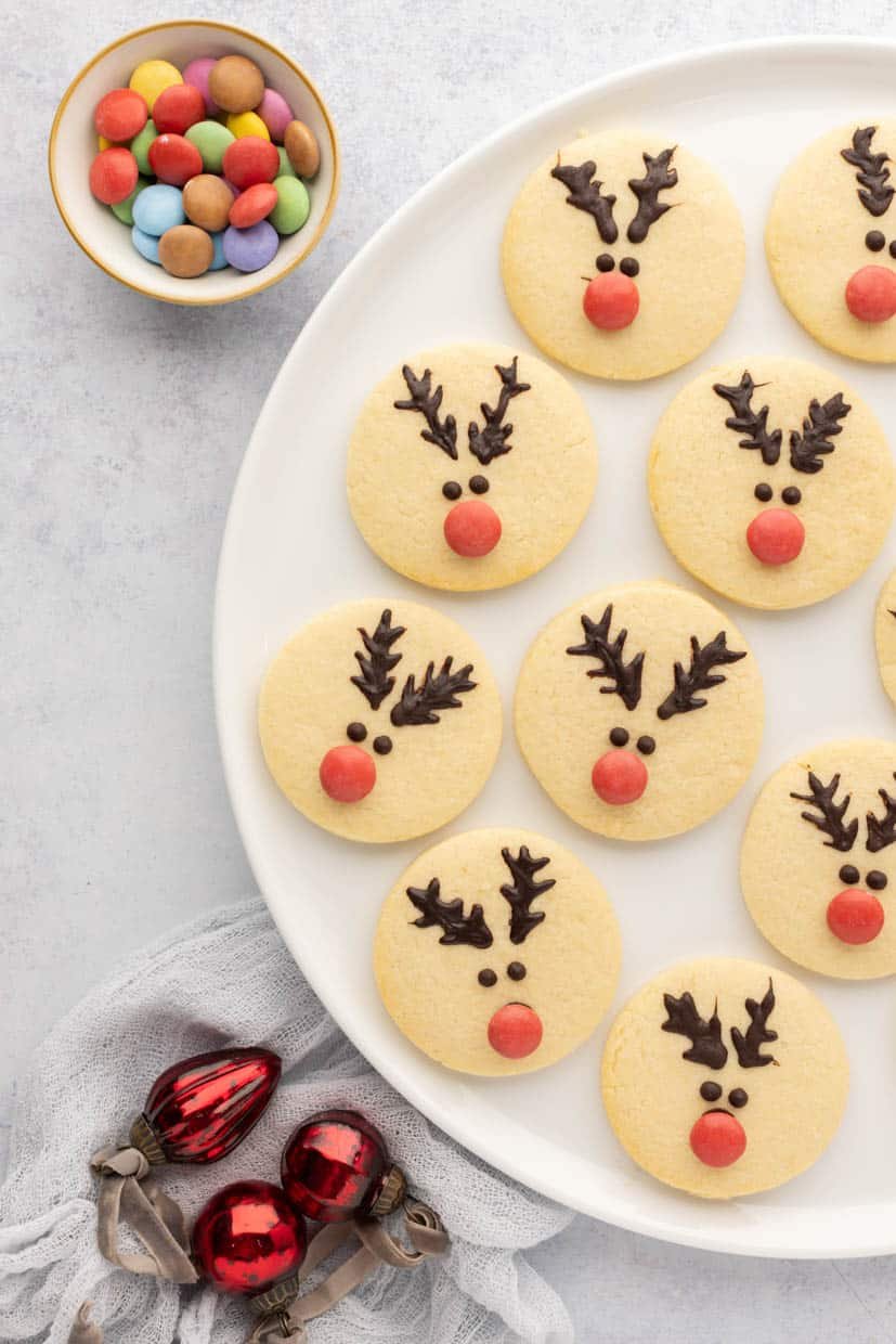 A plate of reindeer-themed cookies with chocolate antlers and red candy noses. Nearby, there is a bowl of colorful candies and red glass ornaments.