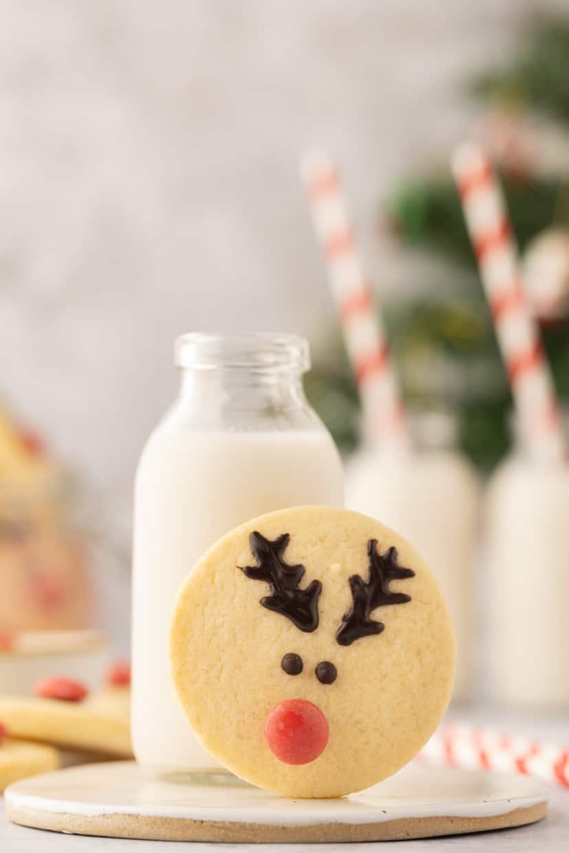 A reindeer-shaped cookie with chocolate antlers and a red nose is propped against a bottle of milk. Striped straws are in the background.