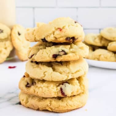 A stack of five cookies with chocolate chips, with more cookies on a plate in the background, set on a white marble countertop.