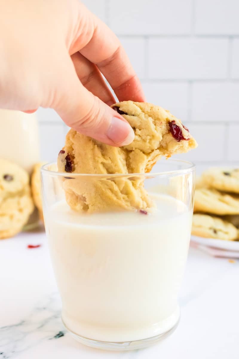 A hand dips a chocolate chip cookie with cranberries into a glass of milk.