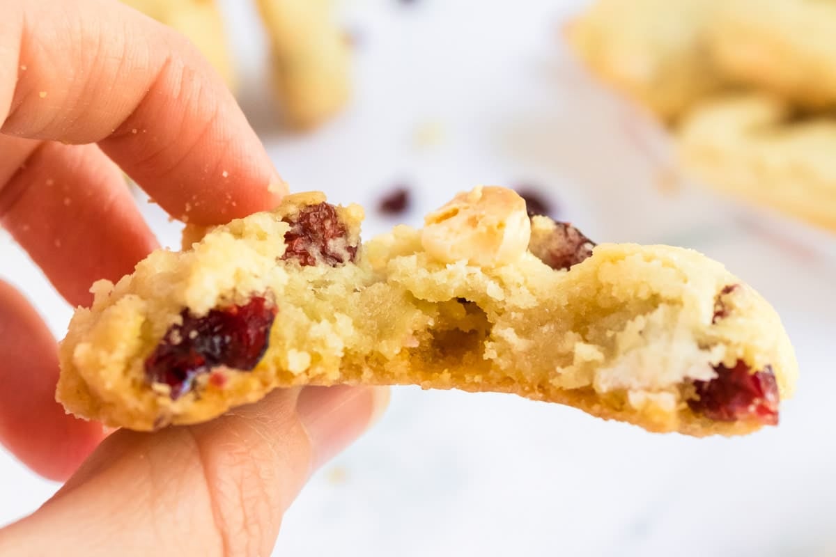 Close-up of a hand holding a bitten cookie with visible cranberries and white chocolate chunks.